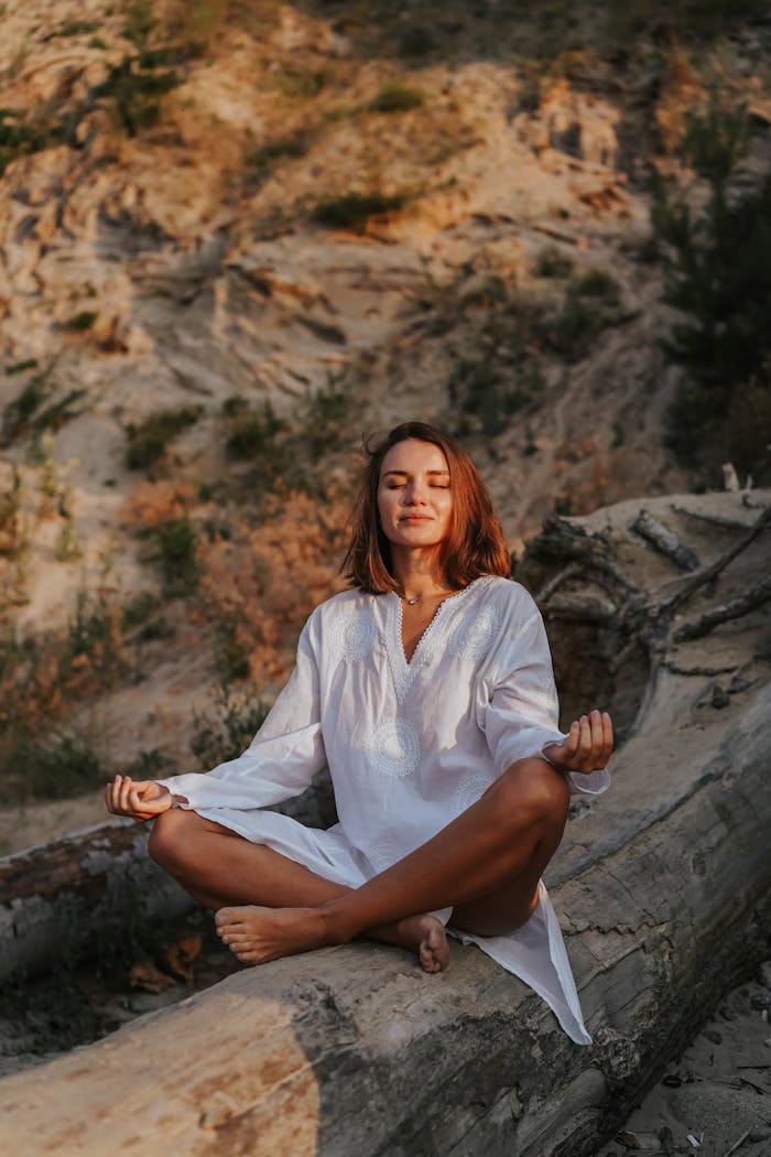 Woman in white dress meditating outdoors on a tree log, embracing tranquility and wellness.